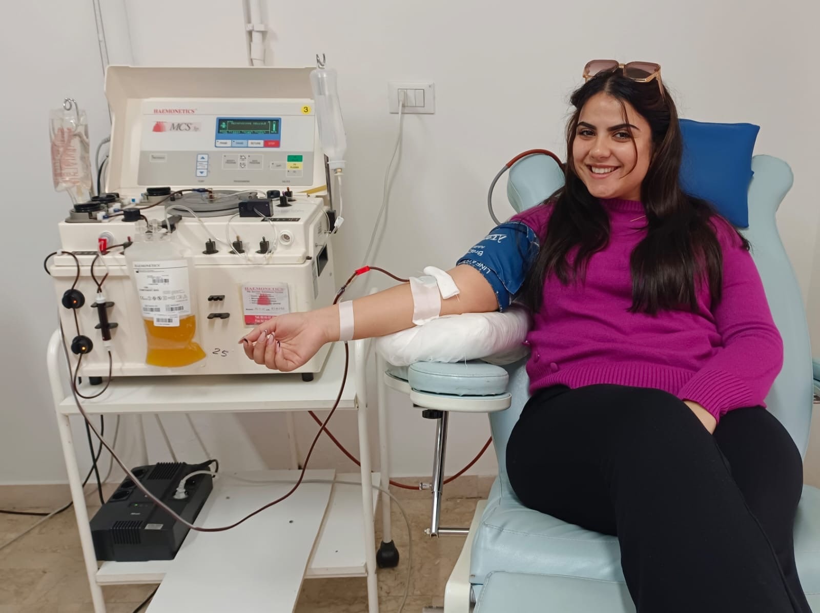 Una donatrice sorridente durante una donazione di plasma da aferesi, con la sacca di plasma e il separatore cellulare accanto alla poltrona